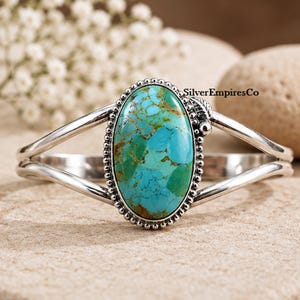 May include: A silver cuff bracelet featuring a large, oval-shaped turquoise stone with brown matrix veining. The stone is set in a beaded silver bezel with a small leaf detail. The band splits into two polished silver arms. Text reads SilverEmpiresCo.