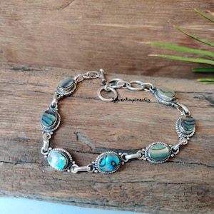 Könnte beinhalten: Ein silbernes Armband mit fünf ovalen Einlagen aus Abalone-Muschel. Das Armband hat einen Schiebeverschluss.