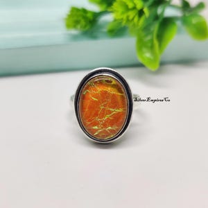 Könnte beinhalten: Ein silberner Ring mit einem ovalen, orangefarbenen Edelstein. Der Edelstein hat eine strukturierte Oberfläche mit grünen und gelben Akzenten. Der Ring befindet sich auf einer weißen Oberfläche mit grünem Laub im Hintergrund. Der Text "Silver Empires Co" ist sichtbar.
