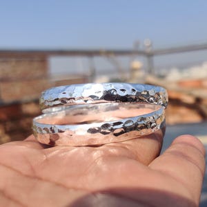 Könnte beinhalten: Zwei silberne, gehämmerte Armreifen. Die Armreifen sind übereinander gestapelt und haben eine strukturierte, gehämmerte Oberfläche.