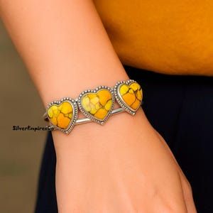 May include: A silver bracelet featuring three heart-shaped stones with yellow and orange patterns. The stones are framed with a dotted silver border. The bracelet is worn on a wrist.