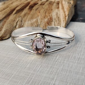 Könnte beinhalten: Silbernes Armreif-Armband mit einem rosafarbenen ovalen Edelstein, der in einer silbernen und goldenen Fassung gefasst ist. Das Armband hat ein zartes Design mit drei dünnen Bändern.