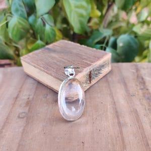 May include: A clear oval-shaped crystal pendant with a silver bail. The pendant is on a wooden surface.
