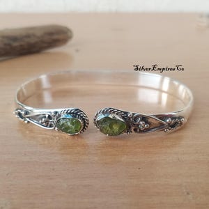 Könnte beinhalten: Offener Armreif mit Silber-Finish. Das Armband hat dekorative Details und zwei olivgrüne Edelsteine. Der Text "Silver Empires Co" ist im Bild zu sehen.