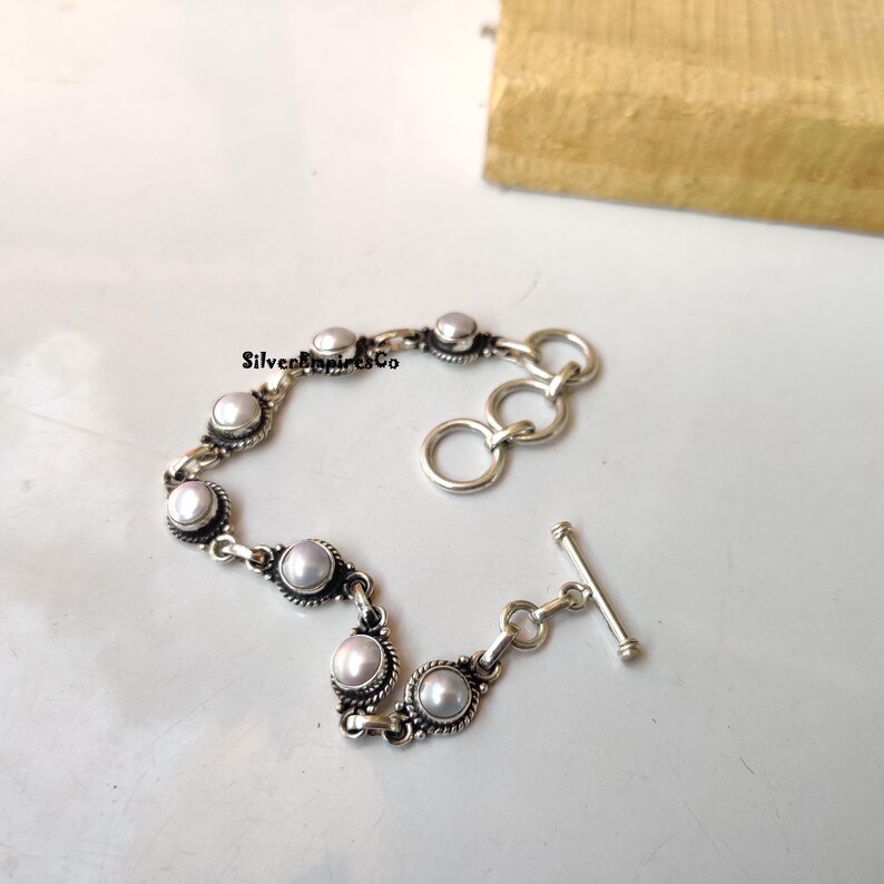 May include: A silver bracelet featuring a series of round, white pearls set in ornate, detailed settings. The bracelet has a toggle clasp and is displayed on a white surface.