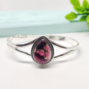 May include: A silver cuff bracelet with a teardrop-shaped, pink and gold stone centerpiece. The stone is framed by a beaded silver border. The bracelet has a double-band design and the text "Silver Empires Co" is visible.