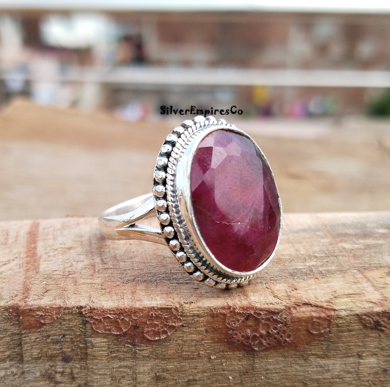 Puede incluir: Un anillo de plata con una gran gema de rub&iacute; ovalada y facetada. El rub&iacute; es de color rojo intenso y est&aacute; engastado en una banda de plata con un borde de cuentas. El anillo se muestra sobre una superficie de madera.
