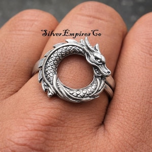 Könnte beinhalten: Ein silberner Ring mit einem Drachendesign. Der Drache umschließt den Finger, Kopf und Schwanz treffen sich. Der Ring weist aufwendige Details wie Schuppen und eine strukturierte Oberfläche auf. Der Text "Silver Empires Co" ist sichtbar.