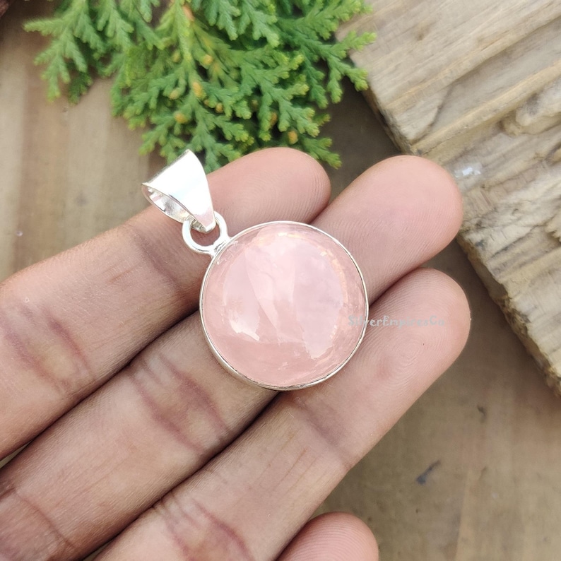 May include: A silver pendant with a round, light pink rose quartz gemstone. The pendant is on a hand.
