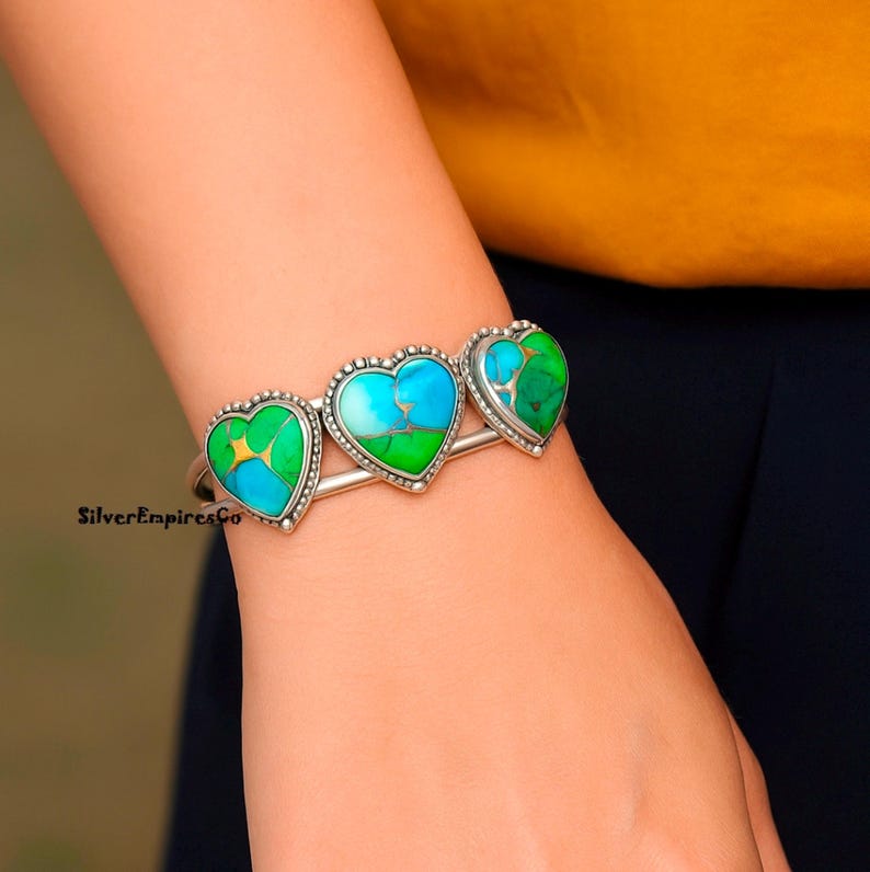 May include: Silver cuff bracelet featuring three heart-shaped turquoise and green stones. The stones are set in a silver frame with a beaded edge. The bracelet is a unique piece of jewelry.