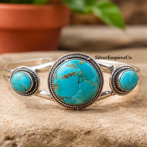 May include: A silver cuff bracelet featuring three round turquoise stones with brown matrix patterns. The central stone is larger than the two side stones, and each is framed by a twisted silver rope border. The text SilverEmpiresCo is visible.