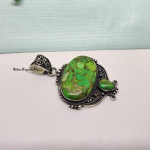 May include: A silver pendant with a large, oval-shaped green stone with brown veining. A smaller, teardrop-shaped green stone is adjacent. The pendant has ornate silver detailing and a decorative bail. The text "Silver Empires Co" is visible.
