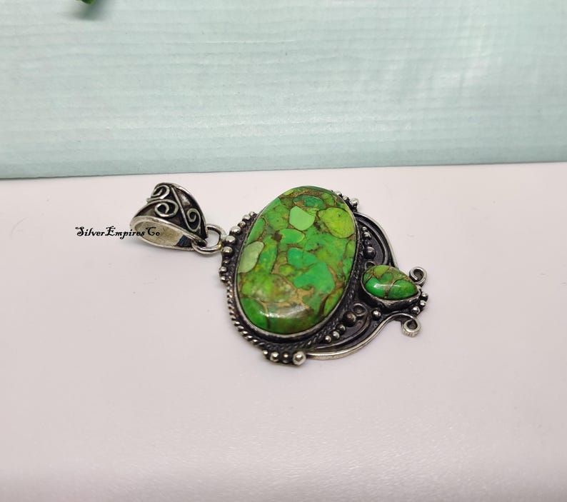 May include: A silver pendant with a large, oval-shaped green stone with brown veining. A smaller, teardrop-shaped green stone is adjacent. The pendant has ornate silver detailing and a decorative bail. The text "Silver Empires Co" is visible.