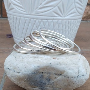 May include: A set of six silver bangles stacked on top of each other. The bangles are smooth and shiny, and they have a slightly rounded shape. The bangles are resting on a gray rock.