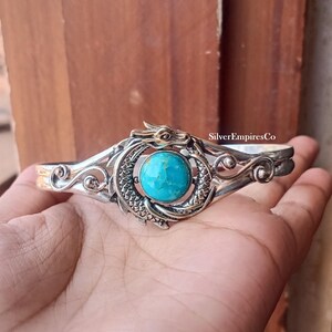 Könnte beinhalten: Ein silbernes Armband mit einem zentralen runden Türkisstein, der von einem detaillierten Drachenmotiv mit Schuppen und Kopf umgeben ist. Das Metall hat eine polierte Oberfläche mit Schnörkeln an den Seiten. Der Text SilverEmpiresCo ist sichtbar.