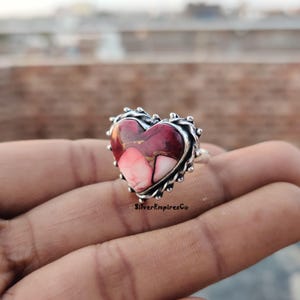 Könnte beinhalten: Herzförmiger Ring mit Silberband und Verbundstein. Der Stein weist eine Mischung aus roten, rosa und weißen Farbtönen auf. Der Ring hat einen strukturierten, perlenbesetzten Rand. Der Text "SilverEmpiresCo" ist sichtbar.