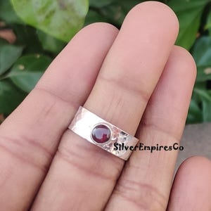 Op de afbeelding: Een zilveren ring met een gehammerde textuur en een rode edelsteen in het midden. De ring zit aan een vinger.