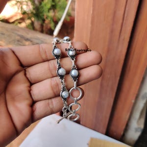 Puede incluir: Una pulsera de plata con perlas blancas engastadas en engastes de metal oscuro y ornamentado. La pulsera tiene un cierre de palanca y se sostiene en una mano. El texto "SilverEmpiresCo" es visible.