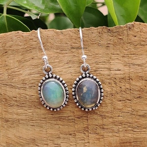 May include: Silver drop earrings featuring oval gemstones. One gemstone has a green hue, the other blue. The earrings are set in a beaded silver frame and hang from a simple hook. The earrings are displayed on a wooden surface.