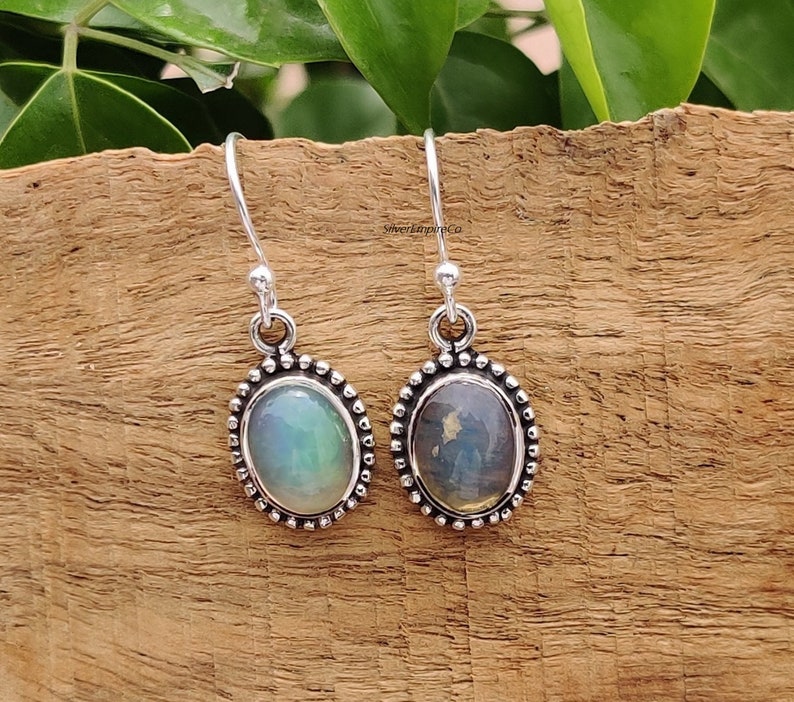 May include: Silver drop earrings featuring oval gemstones. One gemstone has a green hue, the other blue. The earrings are set in a beaded silver frame and hang from a simple hook. The earrings are displayed on a wooden surface.