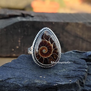 Op de afbeelding: Een zilveren ring met een traanvormige ammoniet edelsteen. De ammoniet is bruin en wit met een spiraalvormig patroon. De ring is gezet in een zilveren band met een touwachtig ontwerp.