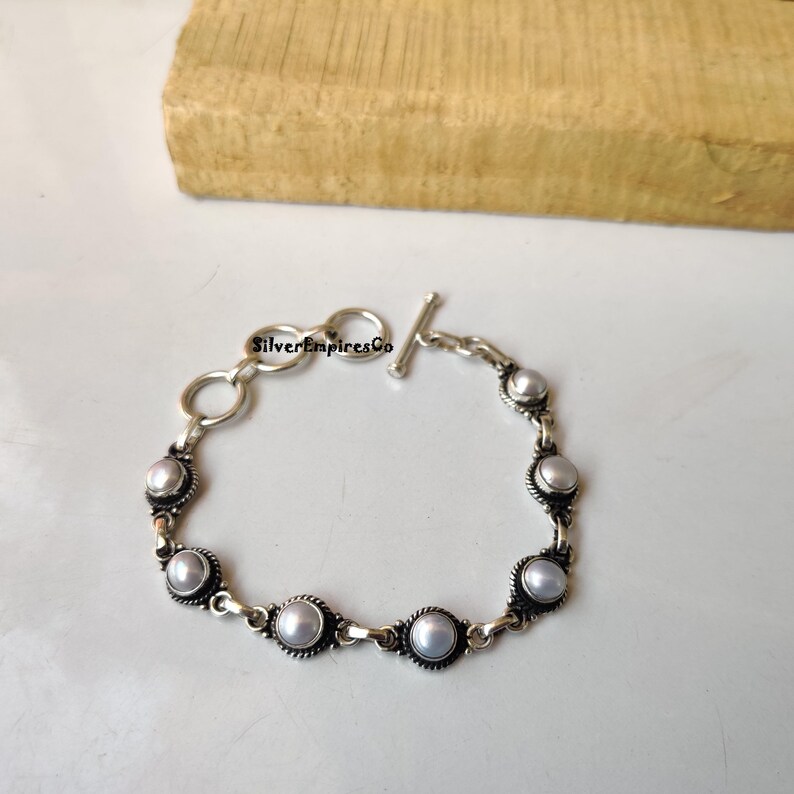 May include: A silver bracelet featuring seven round, light-colored pearls. The bracelet has a toggle clasp and is connected by small, decorative links. The text "SilverEmpiresCo" is visible on the bracelet.
