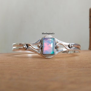 Op de afbeelding: Een zilveren armband met een delicaat ontwerp met een zwierig patroon en een rechthoekige opaal edelsteen in het midden. De opaal heeft een iriserende glans met roze, blauwe en groene tinten.