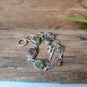 以下が含まれることがあります： 留め金と7つのファセットカットされた宝石が付いたシルバーブレスレット。宝石は緑、ピンク、赤です。ブレスレットは木製の表面にあります。