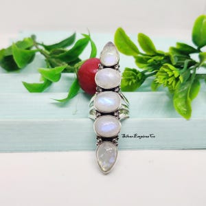 Könnte beinhalten: Silberring mit fünf irisierenden Mondstein-Cabochons in vertikaler Anordnung. Der Ring hat ein detailliertes Silberband. Im Hintergrund sind Grün und eine rote Frucht zu sehen.