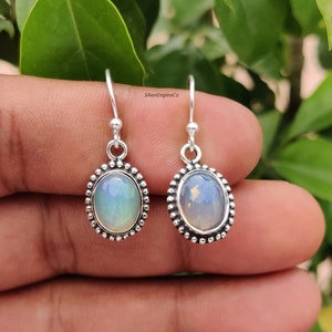 May include: Silver drop earrings with oval opal stones. The opals have a milky, iridescent appearance, set in a beaded silver frame. The earrings are shown against a natural green background.