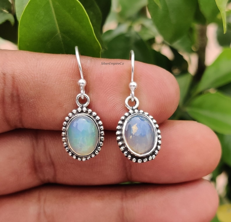 May include: Silver drop earrings with oval opal stones. The opals have a milky, iridescent appearance, set in a beaded silver frame. The earrings are shown against a natural green background.