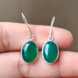 May include: A pair of silver dangle earrings featuring oval-shaped, glossy green gemstones. The gemstones are set in a silver bezel, and the earrings have a simple hook design. The earrings are displayed against a neutral background.