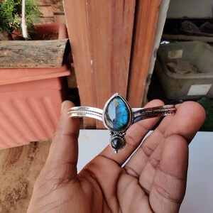 May include: A silver bracelet featuring a teardrop-shaped labradorite gemstone. The gemstone displays a blue and green iridescent sheen. The bracelet has a delicate design with a small silver bead below the main stone.