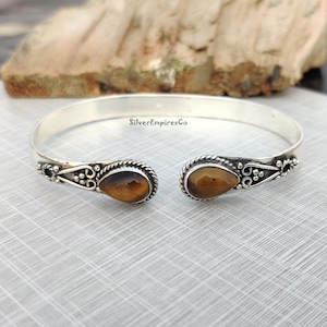 Könnte beinhalten: Silberner Armreif mit zwei tropfenförmigen Tigeraugen-Steinen. Das Armband hat ein zartes, verziertes Design mit kleinen Perlen und Schnörkeln.