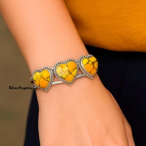 May include: A silver bracelet featuring three heart-shaped stones with yellow and orange patterns. The stones are framed with a dotted silver border. The bracelet is worn on a wrist.