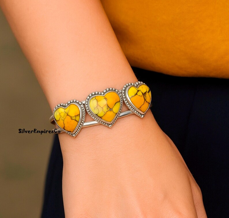 May include: A silver bracelet featuring three heart-shaped stones with yellow and orange patterns. The stones are framed with a dotted silver border. The bracelet is worn on a wrist.