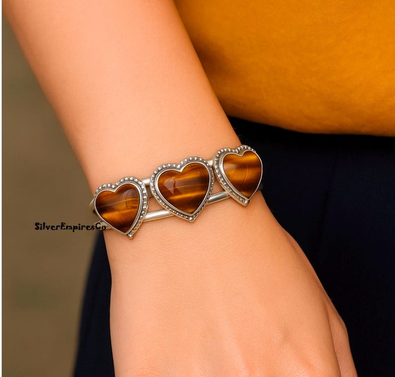 May include: A silver bracelet featuring three heart-shaped tiger's eye stones. The bracelet has a simple, elegant design with a beaded border around each heart. The warm brown and gold tones of the stones contrast with the silver setting.