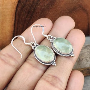 Puede incluir: Un par de pendientes de plata con gotas ovaladas de piedra preciosa verde. Los pendientes tienen un diseño delicado con un borde plateado alrededor de las piedras.