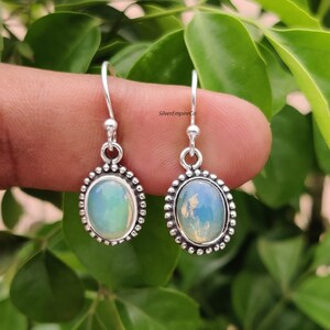 May include: Silver drop earrings featuring oval opal stones. The earrings have a beaded border and a hook fastening. The opal stones display a milky, iridescent quality, with touches of blue and green. The earrings are set against a green backdrop.