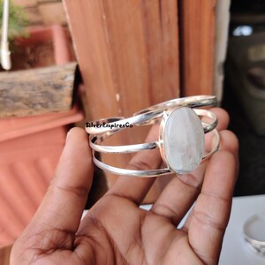 Könnte beinhalten: Ein silbernes Armreif-Armband mit einem großen, ovalen, hellfarbigen Edelstein. Das Armband hat ein Mehrband-Design mit dem Text "SilverEmpiresCo". Der Edelstein ist in einer silbernen Lünette gefasst.