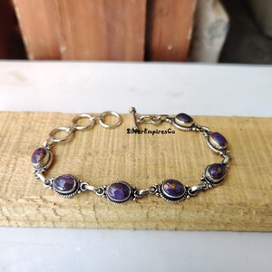 Könnte beinhalten: Ein silbernes Armband mit einem Schiebeverschluss verfügt über ovale, violette und goldene Steine. Das Armband liegt auf einer Holzoberfläche. "SilverEmpiresCo" ist auf der Oberfläche aufgedruckt.