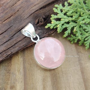 May include: A silver pendant with a round, light pink rose quartz gemstone. The pendant is on a wooden surface.