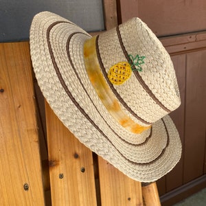 May include: A straw hat with a wide brim and a decorative pineapple. The hat features brown stripes and a yellow and orange ribbon around the crown. The pineapple is yellow with green leaves.