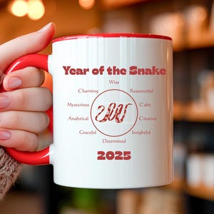 May include: White ceramic mug with a red handle and interior. The mug features the text "Year of the Snake" and a stylized snake design, along with the year "2025". Additional words include "Wise", "Charming", "Mysterious", "Analytical", "Graceful", "Resourceful", "Calm", "Creative", "Insightful", and "Determined".