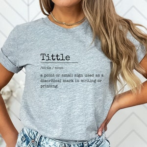May include: A gray t-shirt with the word "Tittle" defined as a "point or small sign used as a diacritical mark in writing or printing."