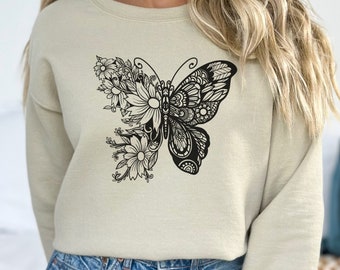 May include: Sand-colored sweatshirt featuring a black butterfly design. The butterfly is split, with one side floral and the other a detailed pattern. The text "ELM Tees & Apparel" is at the bottom left. The word "SAND" is in a circle at the bottom right.