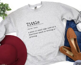 May include: A gray sweatshirt with the word "Tittle" and its definition printed in black. The definition is: "tit·tle / noun a point or small sign used as a diacritical mark in writing or printing."