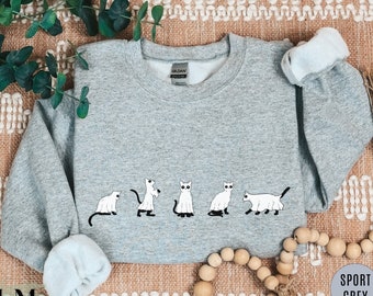 Puede incluir: Sudadera gris deportiva con cuello redondo y mangas largas. Presenta una fila de gatos de dibujos animados en diferentes poses. Los gatos son blancos con detalles negros. La marca "Gildan" es visible.