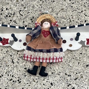 以下が含まれることがあります： 人形のフィギュアが付いた装飾的な壁棚。棚は白で、黒と白のストライプのアクセントと黒いフックが付いています。人形は麦わら帽子、チェック柄のジャケット、ハートのアップリケが付いたドレスを着ています。ボタンと星型の生地のアクセントも付いています。
