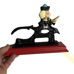 Puede incluir: Un cascanueces vintage de hierro fundido con forma de soldado. La figura tiene cabello rubio, un sombrero azul y un uniforme negro con detalles dorados. Está montado sobre una base de madera roja.